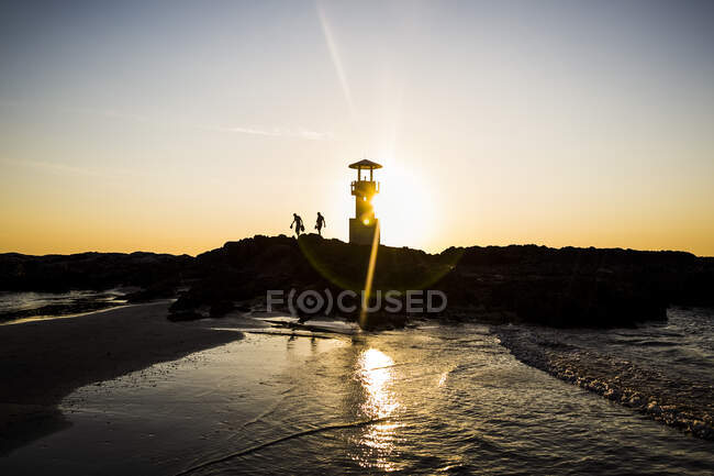 Silhouettes of two people walking past lighthouse by the ocean at sunset. — Stock Photo