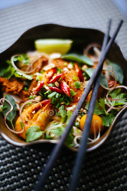 Hohe Nahaufnahme von Essstäbchen auf Schüssel mit asiatischem Essen mit Nudeln, Garnelen, Gemüse und Chiligarnitur. — Stockfoto