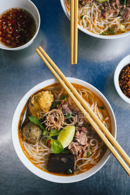 High angle close-up of bowls of Bun Bo Hue with beef, crab balls, blood sausage and herbs. — Stock Photo