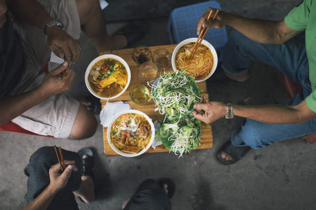 High angle close-up of three men tucking into bowls of noodles for breakfast in a small alleyway. — Stock Photo