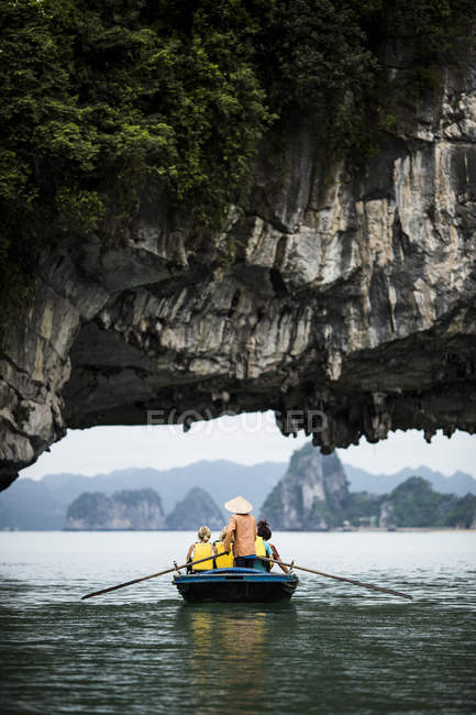 Rear view of man wearing straw hat transporting small group of people on a boat, rowing underneath natural rock arch, Bai Tu Long, Vietnam. — Stock Photo