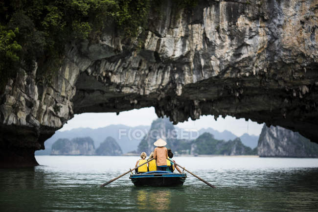 Vue arrière de l'homme portant un chapeau de paille transportant un petit groupe de personnes sur un bateau, ramant sous une arche de roche naturelle, Bai Tu Long, Vietnam . — Photo de stock