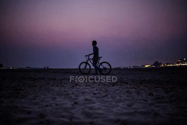 Silhouette of boy pushing bicycle on bank of a river at sunset in Laos — Stock Photo