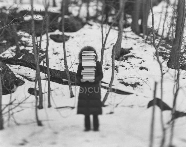 Child wearing a coat standing in winter forest, holding tall stack of books. — Stock Photo