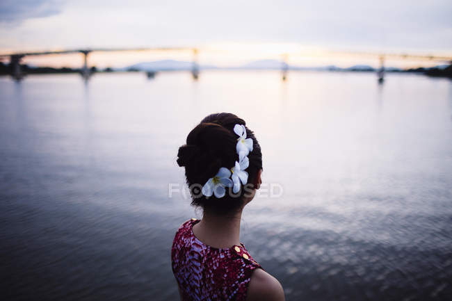 Vue arrière de la femme avec des fleurs dans les cheveux, debout au bord de la mer au coucher du soleil, pont au loin . — Photo de stock