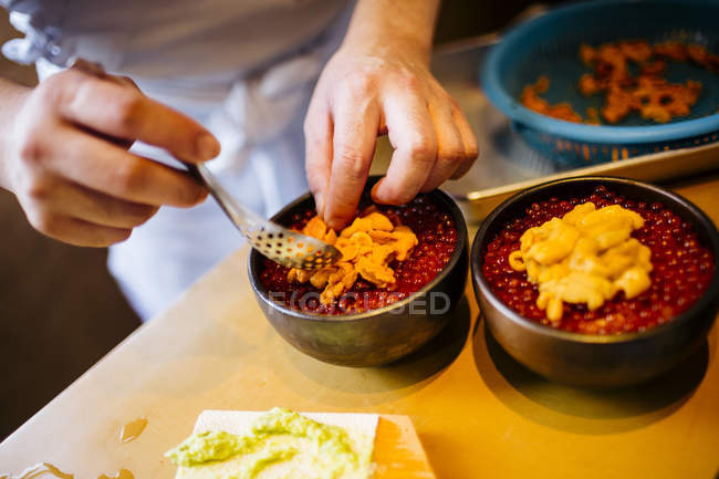 Alto ângulo close-up de chef preparando ouriço do mar com ovas de salmão e tigelas de arroz . — Fotografia de Stock
