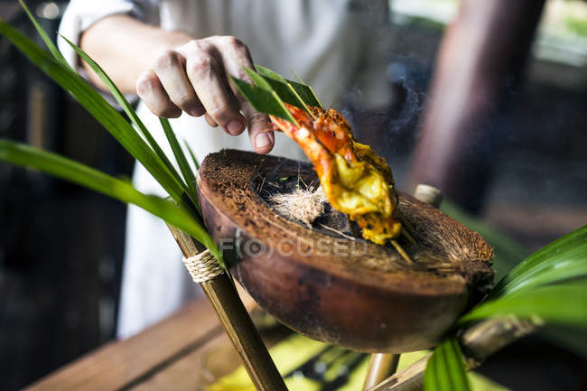 Close-up of chef preparing charcoal-grilled prawn satay on a coconut shell. — Stock Photo