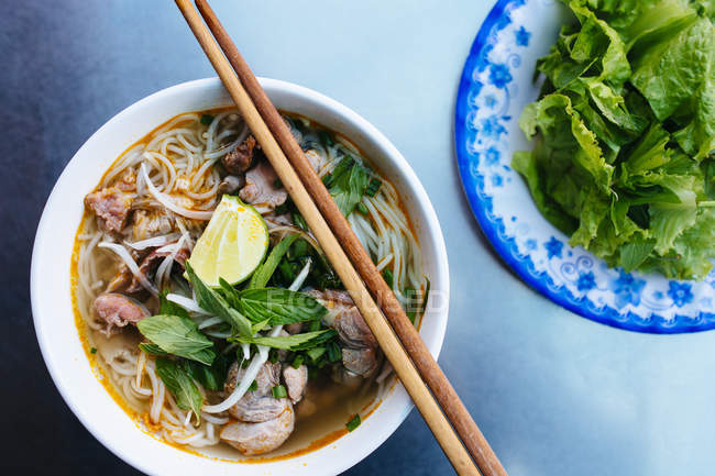 High angle close-up of a bowl of bun bo hue beef noodle soup and plate with green salad. — Stock Photo