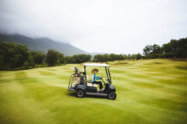 Frau mit traditionellem Strohhut fährt Golfcart auf grünem Gras eines Golfplatzes unter bewölktem Himmel. — Stockfoto
