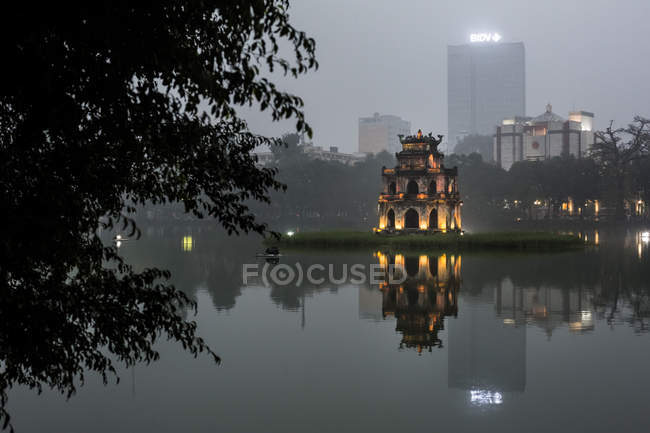 Exterior of illuminated temple reflected in lake at dusk, skyscrapers in distance, Hanoi, Vietnam. — Stock Photo