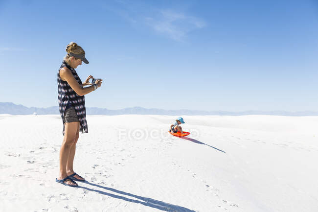 Mère photographiant son fils en traîneau sur des dunes de sable — Photo de stock