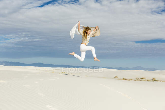 Niña de 13 años bailando y saltando en medio del aire en el espacio abierto sobre dunas de arena blanca. - foto de stock