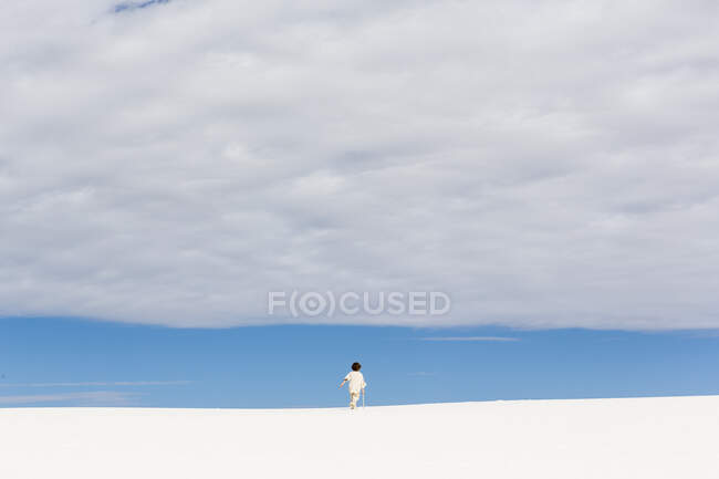 Niño de 6 años caminando en dunas de arena blanca, una tira de cielo azul en el horizonte y nubes gruesas. - foto de stock