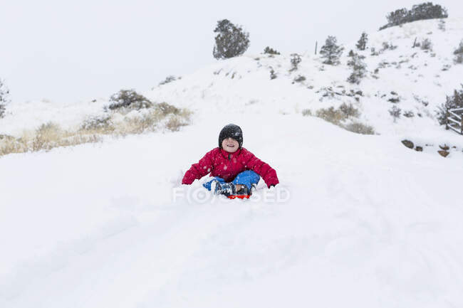 Симпатичный счастливый мальчик катается на санках по склону зимой — стоковое фото