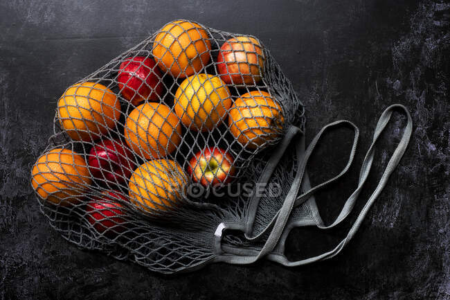 High angle close up of red apples and oranges in grey net bag on black background. — Stock Photo