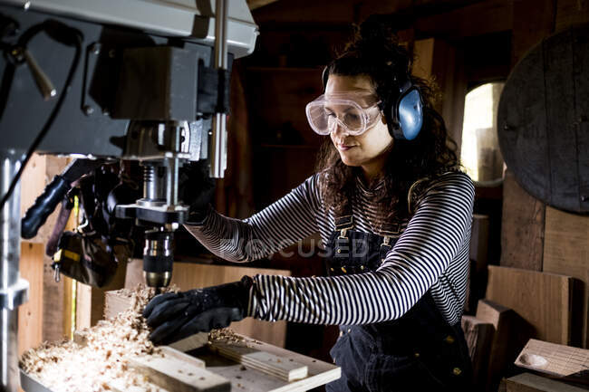 Mulher com cabelos castanhos longos vestindo dungarees, óculos de segurança e protetores de ouvido em pé na oficina de madeira, usando broca elétrica. — Fotografia de Stock