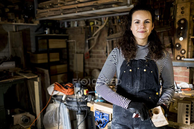 Woman with long brown hair wearing dungarees standing in wood workshop, smiling at camera. — Stock Photo