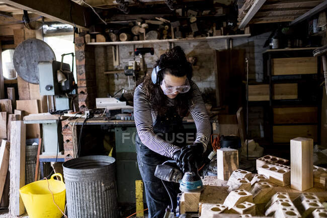 Frau mit langen braunen Haaren mit Latzhose, Schutzbrille und Ohrenschutz steht in Holzwerkstatt und benutzt Schleifer. — Stockfoto