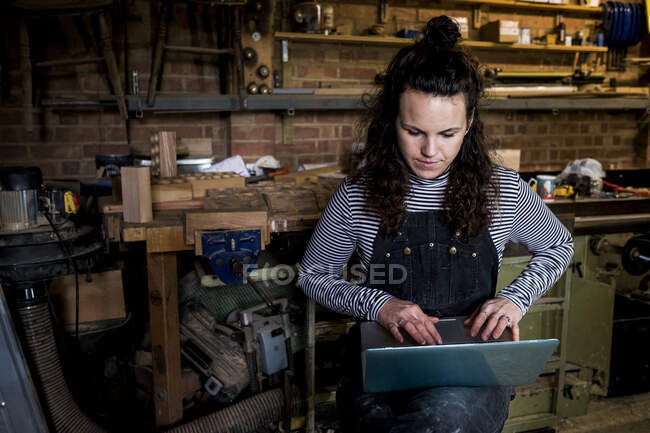 Frau mit langen braunen Haaren in Latzhose steht in Holzwerkstatt und tippt auf Laptop. — Stockfoto