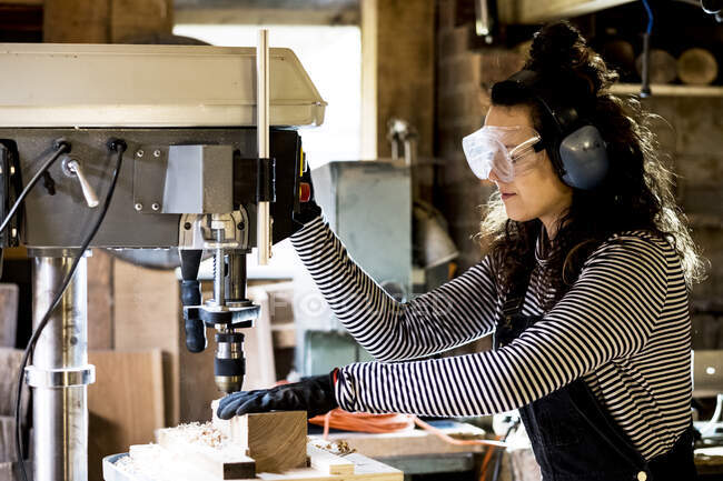 Woman with long brown hair wearing dungarees, safety glasses and ear protectors standing in wood workshop, using electric drill. — Stock Photo