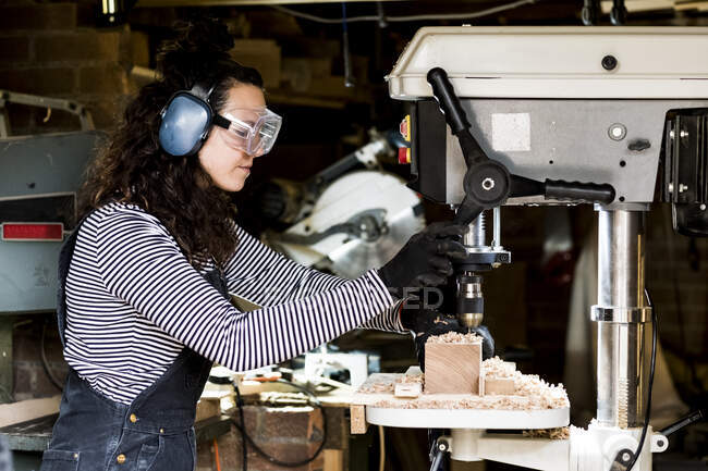Donna con lunghi capelli castani che indossano unghie, occhiali di sicurezza e protezioni dell'orecchio in piedi in officina di legno, utilizzando trapano elettrico. — Foto stock