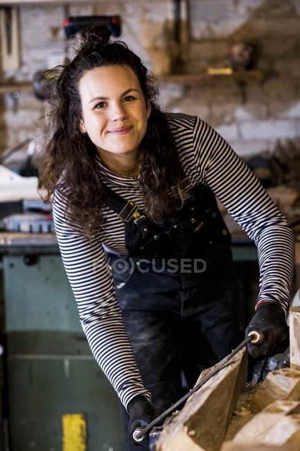 Woman with long brown hair wearing dungarees standing in wood workshop, using plane on piece of wood. — Stock Photo