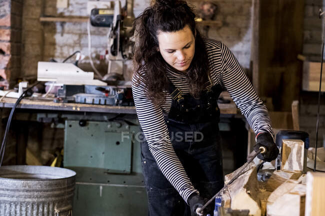 Frau mit langen braunen Haaren in Latzhose steht in Holzwerkstatt und benutzt Hobel auf Stück Holz. — Stockfoto