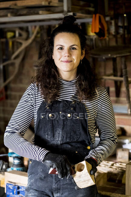 Woman with long brown hair wearing dungarees standing in wood workshop, smiling at camera. — Stock Photo