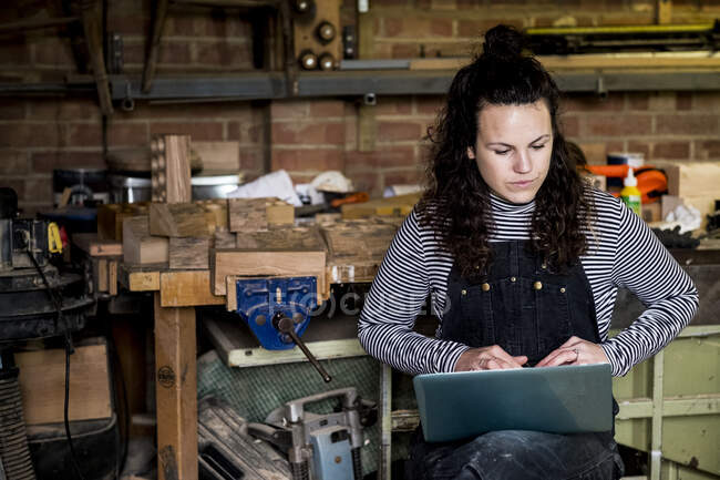 Mujer con pelo marrón largo que lleva calabazas de pie en el taller de madera, escribiendo en ordenador portátil.. - foto de stock