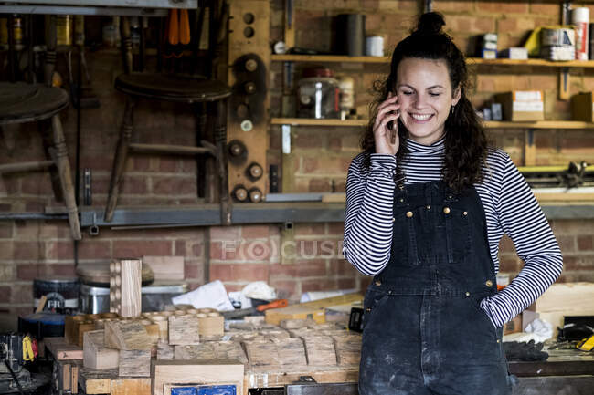 Frau mit langen braunen Haaren in Latzhose steht in Holzwerkstatt und telefoniert mit Handy. — Stockfoto