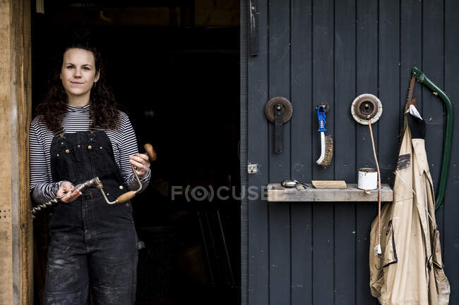 Woman with long brown hair wearing dungarees standing at entrance to wood workshop, looking at camera. — Stock Photo