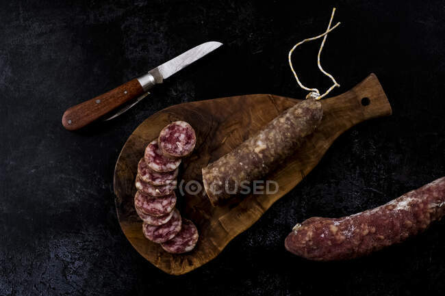 High angle close up of knife, sliced salami on wooden cutting board on black background. — Stock Photo