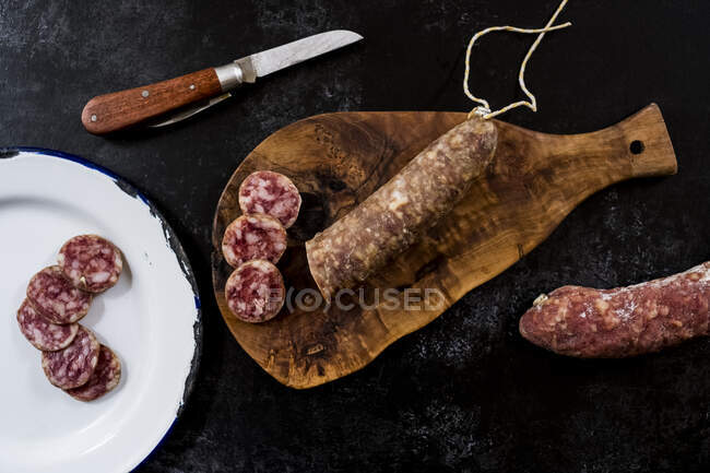 High angle close up of knife, sliced salami on wooden cutting board and white enamel plate on black background. — Stock Photo