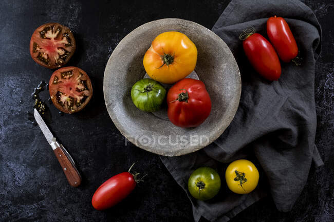Alto ângulo de perto de faca, placa cinza e pano e uma seleção de tomates frescos em fundo preto . — Fotografia de Stock