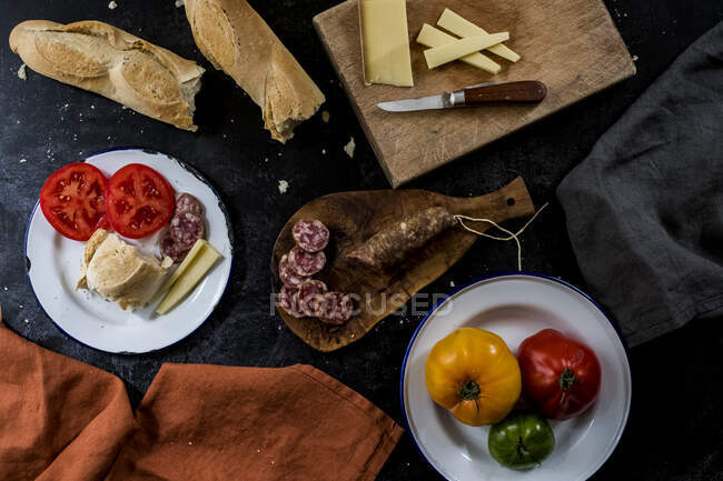Alto ángulo de cierre de una selección de queso, tomates, salami y baguette francesa en placas de esmalte blanco sobre fondo negro . - foto de stock