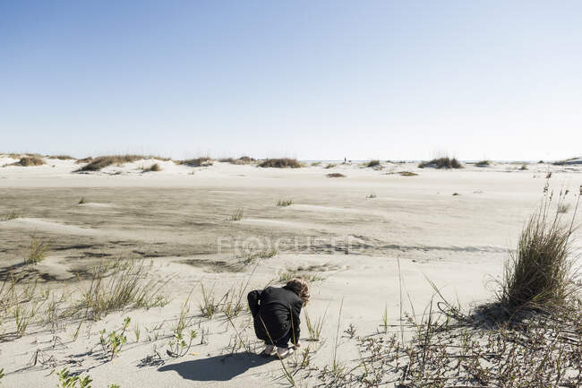 Ragazzo di sei anni in morbide dune di sabbia bianca, piegandosi verso il basso per ispezionare qualcosa. — Foto stock