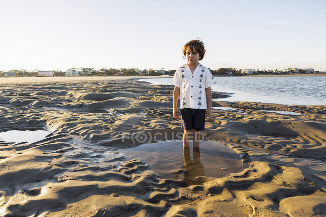 Um menino de seis anos de idade em pé em uma piscina rasa na areia — Fotografia de Stock