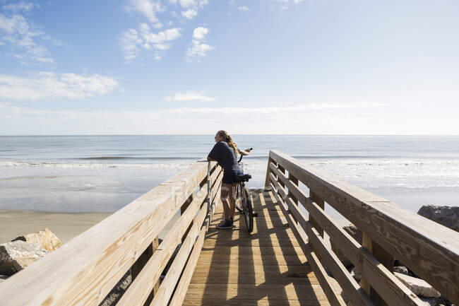 Adolescente menina na ponte de madeira pela praia, com sua bicicleta — Fotografia de Stock