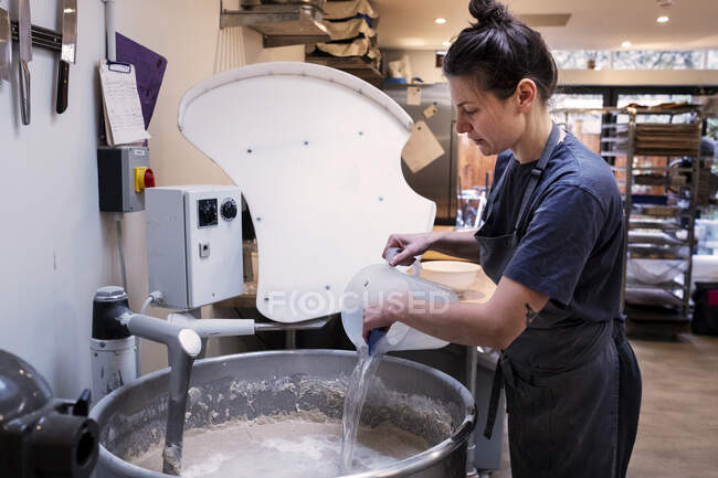 Femme portant un tablier debout dans une boulangerie artisanale, versant de l'eau dans un mélangeur industriel . — Photo de stock