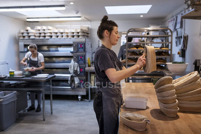 Donna che indossa grembiule in piedi in una panetteria artigianale, preparando cestini di prova . — Foto stock