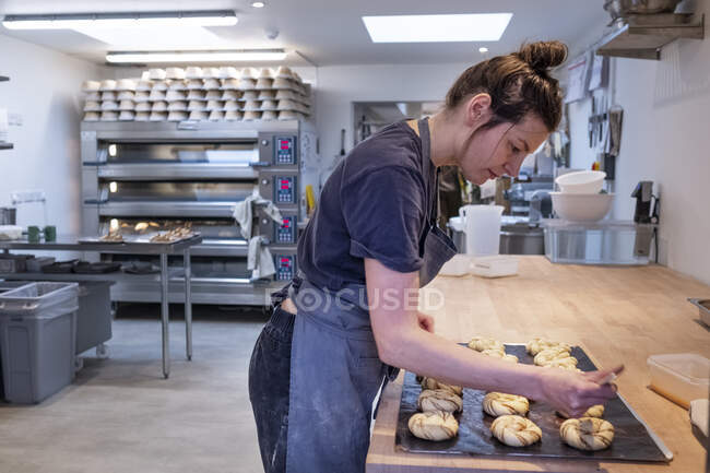 Frau mit Schürze steht in einer Bäckerei und pinselt Zimtbrötchen auf ein Backblech. — Stockfoto