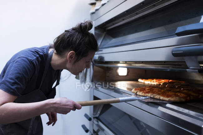 Frau mit Schürze steht in Bäckerei und legt Pizza in Ofen. — Stockfoto