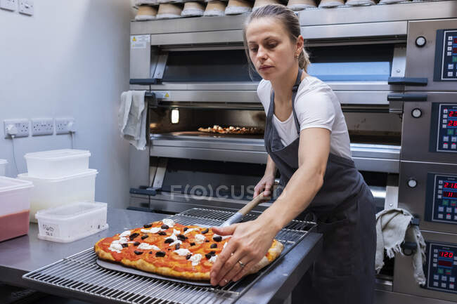 Frau mit Schürze steht in einer Bäckerei und bereitet Pizza für den Ofen zu. — Stockfoto