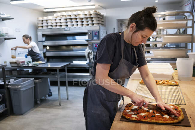 Femme portant un tablier debout dans une boulangerie artisanale, tranchant une pizza fraîchement cuite . — Photo de stock