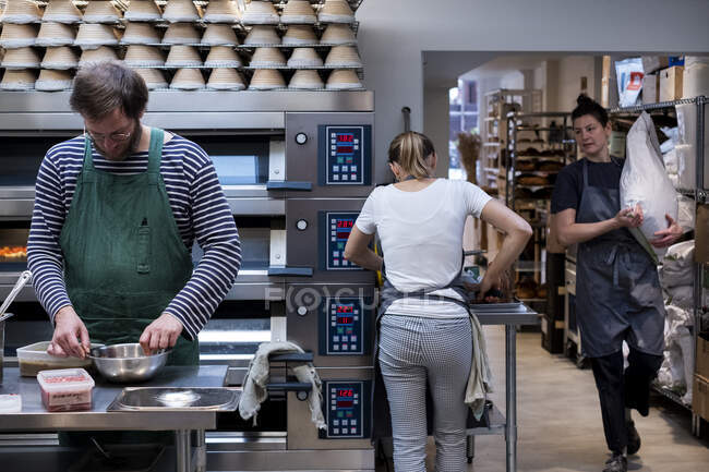 Dos mujeres y un hombre con delantales trabajando en una panadería artesanal . - foto de stock