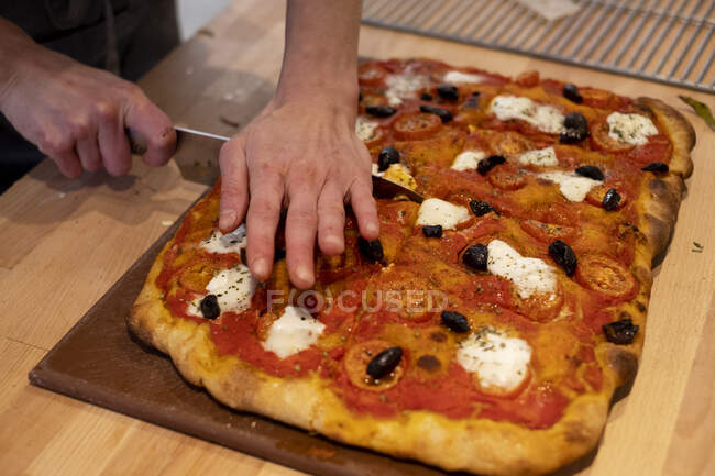 Großaufnahme einer Person, die in einer handwerklichen Bäckerei frisch gebackene Pizza schneidet. — Stockfoto