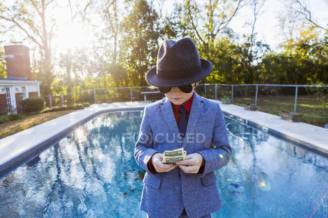 Junge im Anzug und dunkler Brille steht am Rand des Wassers und zählt Dollarscheine.. — Stockfoto