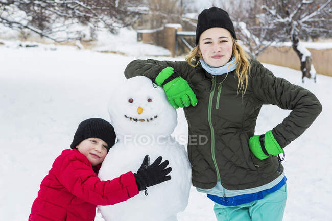 Hermano y hermana, un joven y adolescente que se apoya en muñeco de nieve en invierno - foto de stock