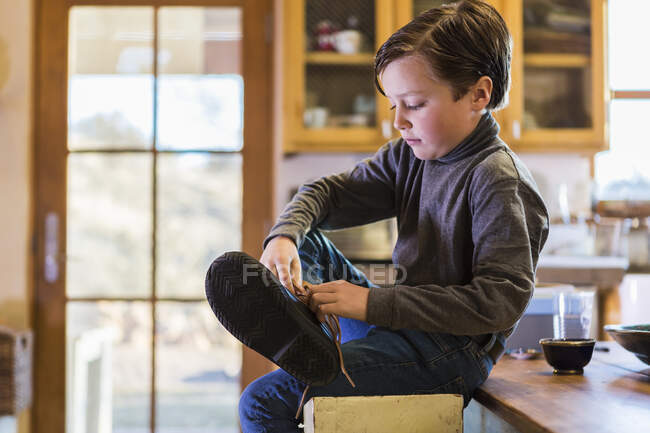 Niño de seis años poniéndose las botas, sentado en un taburete alto . - foto de stock