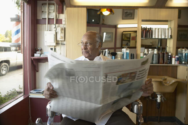 Homem sênior sentado em cadeira de barbeiro com jornal — Fotografia de Stock